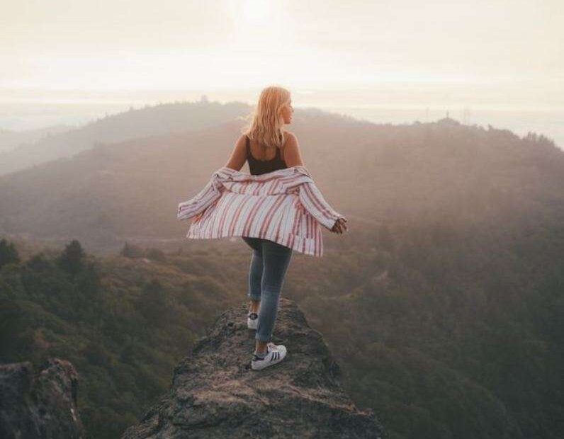 A woman standing confidently on a rocky mountain peak at dawn, looking out over a misty valley with her arms open for building self worth.